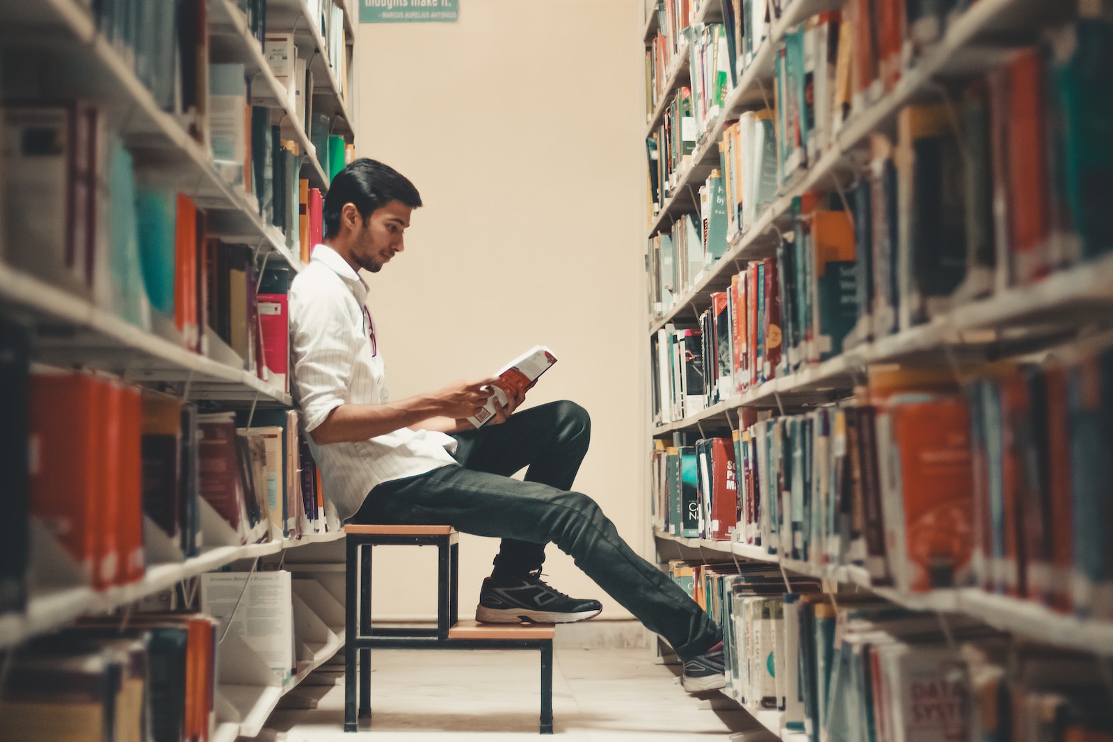 man sitting and leaning near bookshelf while reading book